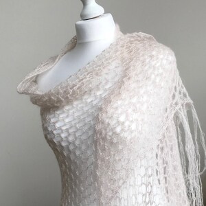 May include: A delicate, off-white crochet shawl with a lacy, open-weave pattern. The shawl is draped over a white mannequin and has a fringed edge.