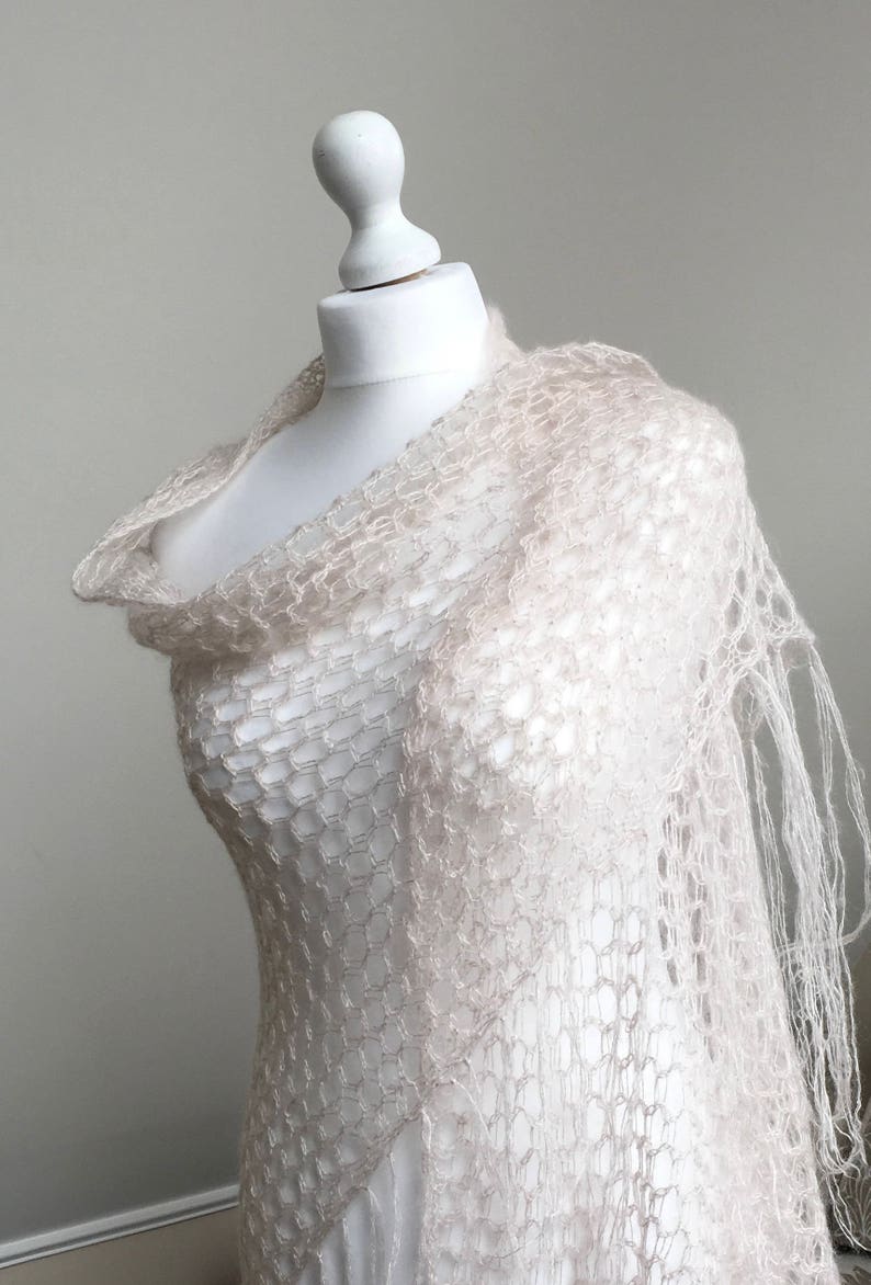 May include: A delicate, off-white crochet shawl with a lacy, open-weave pattern. The shawl is draped over a white mannequin and has a fringed edge.