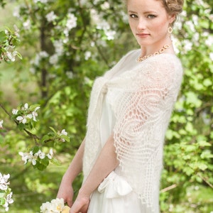 May include: A woman wearing a white lace shawl and a white dress, holding a bouquet of flowers in front of a background of blooming white flowers.