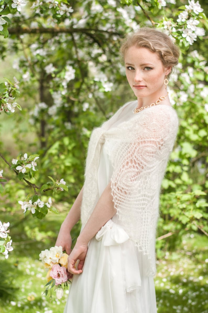 May include: A woman wearing a white lace shawl and a white dress, holding a bouquet of flowers in front of a background of blooming white flowers.