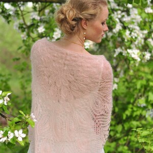 Puede incluir: Una mujer lleva una delicada chal de encaje rosa con un vestido blanco. La chal tiene un delicado patr&oacute;n floral y est&aacute; hecha de una tela suave y ligera. La mujer est&aacute; de pie frente a un fondo de hojas verdes y flores blancas.