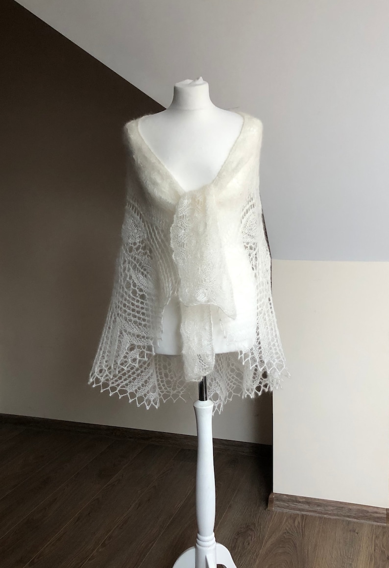 May include: A white knitted shawl with a delicate lace pattern. The shawl is draped over a white mannequin.