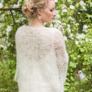 May include: A white lace shawl with a delicate floral pattern. The shawl is draped over the shoulders of a person standing in front of a green background with white flowers.