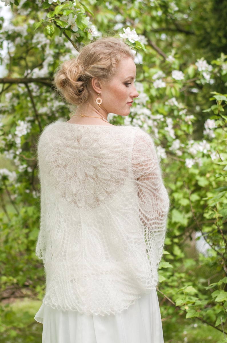 May include: A white lace shawl with a delicate floral pattern. The shawl is draped over the shoulders of a person standing in front of a green background with white flowers.