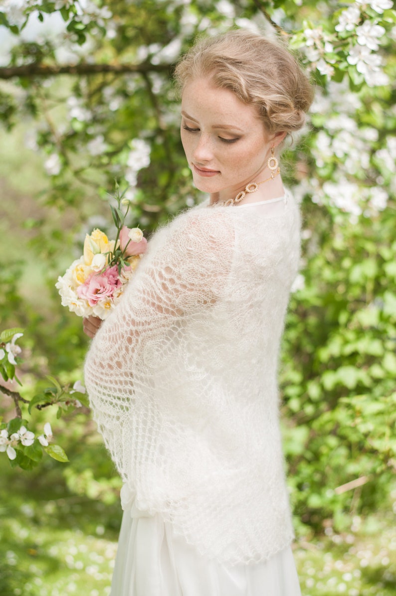 May include: A bride wearing a white lace shawl and holding a bouquet of pink and yellow flowers. She is standing in a garden with green foliage and white flowers.