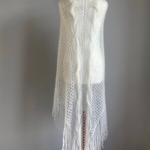 May include: White knitted shawl with a delicate openwork pattern and fringe. The shawl is draped over a white mannequin.