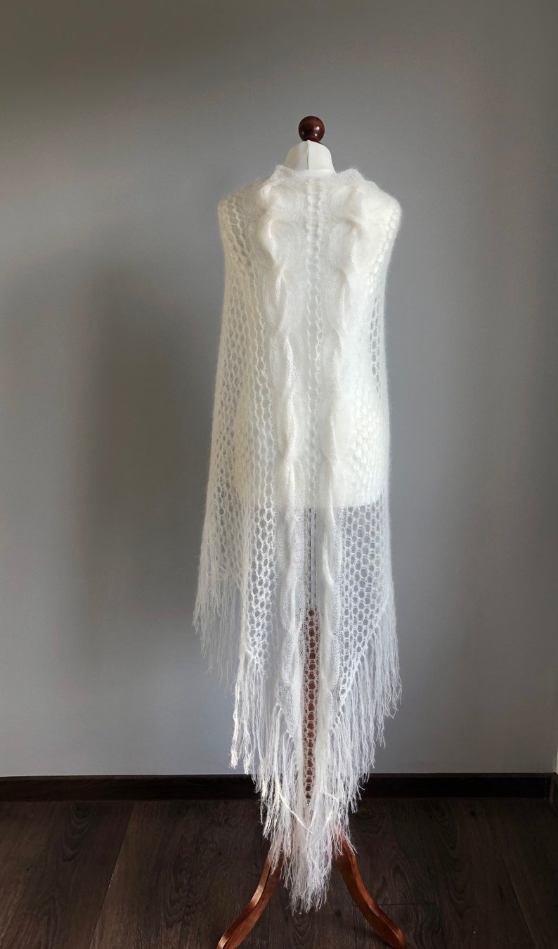 May include: White knitted shawl with a delicate openwork pattern and fringe. The shawl is draped over a white mannequin.