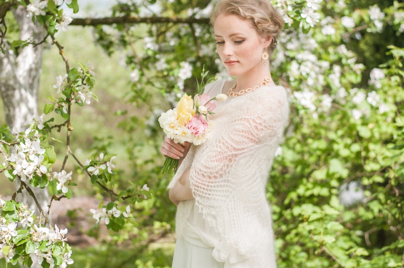 May include: A woman in a white lace shawl holds a bouquet of yellow and pink flowers in front of a blooming tree.
