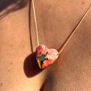 May include: A heart-shaped pendant necklace. The pendant is painted with red, green, yellow, and black colors. The necklace cord is a light tan color. The necklace is worn around the neck.
