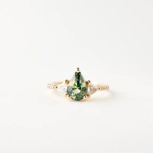 May include: A gold ring with a pear-shaped green gemstone as the centerpiece, flanked by two smaller white triangular stones. The band is adorned with small, sparkling accent stones, creating a delicate and elegant design.