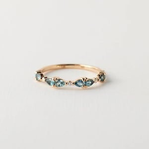 May include: A gold band ring featuring alternating marquise-cut blue gemstones and small round diamonds. The ring is set against a white background, highlighting the ring's delicate design and the sparkle of the gemstones.