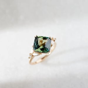 May include: A gold ring with a large, square-cut green gemstone with a hint of yellow. The ring is embellished with smaller, clear gemstones and has a delicate, gold band. The design is elegant and eye-catching.
