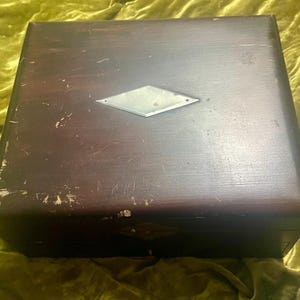 May include: A dark brown wooden box with a diamond-shaped metal accent on the lid. The box has a square shape and shows signs of wear, with scratches and imperfections on the surface. The box is sitting on a green surface.