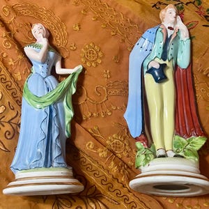 May include: Two ceramic figurines of a man and woman in vintage attire. The woman wears a light blue dress with a green scarf. The man is in a blue coat and red cape, holding a top hat. Both figures stand on white bases.