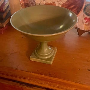 Puede incluir: Un cuenco verde de cerámica sobre pedestal, con una base ancha y poco profunda. El cuenco se asienta sobre una base cuadrada conectada por un tallo redondeado. El cuenco es de color verde apagado y parece ser vintage.