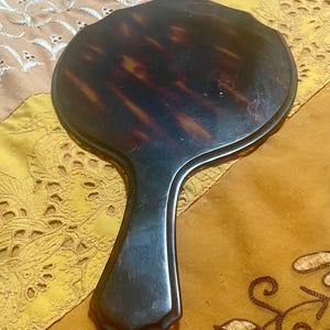 Può includere: Un'antica specchiera a mano con uno specchio ovale scuro a fantasia tartarugata e un lungo manico scuro. Lo specchio è posto su uno sfondo di tessuto fantasia nei toni dell'oro e del beige.