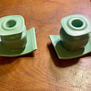 May include: Two matching green ceramic candle holders. Each holder has a square top with a circular opening, sitting atop a rounded base and a square, curved plate. The candle holders are a light green color.