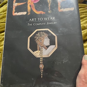 May include: A black hardcover book titled ERTE ART TO WEAR THE COMPLETE JEWELRY. The cover features a large, stylized illustration of a person in profile wearing an ornate, patterned headpiece and long dangling jewelry inside an octagon frame.
