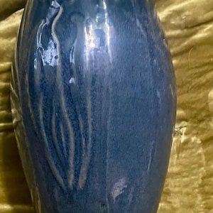 May include: A cylindrical blue ceramic vase with a glossy finish and a raised, textured relief pattern featuring vertical, flowing lines that extend from the top toward the base, creating a subtle decorative effect on the exterior surface.
