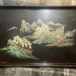 以下が含まれることがあります： 手描きの風景画が描かれた長方形の黒いサービングトレイ。アートワークは、緑、金、赤の色合いの山、木、建物を示しています。トレイには濃い茶色の縁があります。