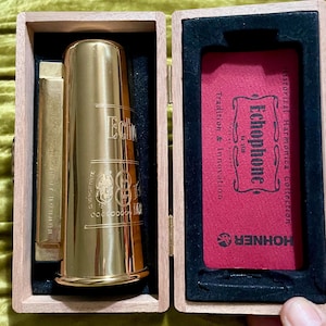 May include: A gold-coloured metal Echophone harmonica housed in a light-coloured wooden box with black velvet lining. The right side of the box contains a red insert with the text Echophone No. 3510 Historical Harmonica Collection Hohner.