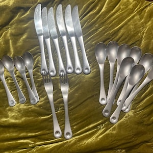 May include: A collection of vintage silverware, including knives, spoons, and forks, arranged on a textured, olive-green surface. The silverware has a matte, silver finish. The spoons have a rounded bowl shape, and the forks have three tines.