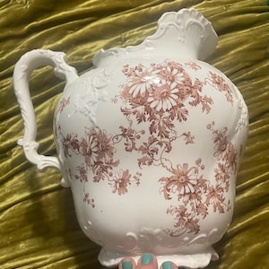 Może przedstawiać: Biały ceramiczny dzban z dużym uchwytem i ozdobnym wylewem. Dzban jest zdobiony kwiatowym wzorem w odcieniach brązu. Dzban ma pękaty korpus i ozdobny uchwyt.