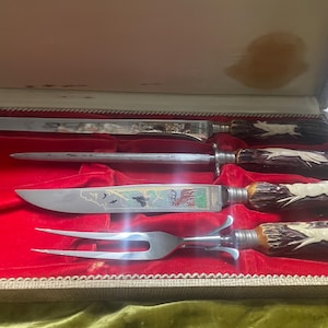 May include: A vintage carving set in a box with a red velvet interior. The set includes a carving knife, a sharpening steel, a carving knife, and a fork. The handles are made of brown material with a white deer design.