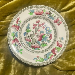 Op de afbeelding: Een ronde keramische bord met een centrale illustratie van een bloeiende boom met roze, blauwe en groene bloesems. De rand is versierd met een zwart geometrisch Grieks sleutelpatroon en extra bloemmotieven in roze en groene tinten.