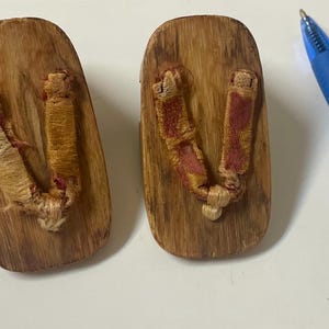 May include: Two wooden geta sandals with brown wood bases and woven straps. The straps are tan and red. A blue pen is in the background.