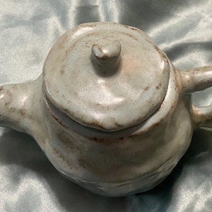 May include: A small, handmade ceramic teapot with a lid. The teapot is a light grey colour with brown accents and a rounded shape. It has a handle and a spout. The teapot is likely used for brewing tea.
