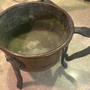 May include: A copper-colored metal bowl with a handle and three legs. The bowl has a dark patina and a hammered handle. The legs are attached to the bowl with rivets. The bowl is likely an antique or vintage item.
