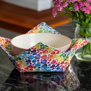 May include: A ceramic bowl sits within a fabric bowl cosy, decorated with a pattern of rainbow-coloured watercolour hearts. A glass jar with pink flowers is visible in the background.