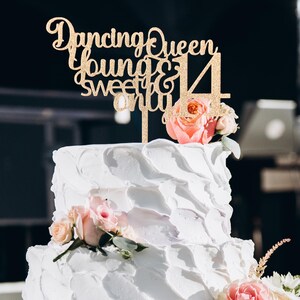 May include: A two-tiered white frosted cake decorated with pink roses and a gold glitter cake topper. The cake topper reads "Dancing Queen, Young & Sweet & 14". The cake is likely for a birthday celebration.