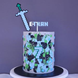 Könnte beinhalten: Eine Geburtstagstorte, verziert mit einem pixeligen Schwert-Tortentopper und dem Namen "ETHAN". Der Kuchen ist mit blauen, grünen und schwarzen Pinselstrichen und quadratischen Akzenten verziert. Eine große "7" ist auf der Vorderseite zu sehen.