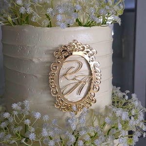 Pode incluir: Um bolo branco em camadas decorado com flores brancas e um monograma dourado. O monograma dourado está em uma moldura oval ornamentada. O bolo tem uma cobertura de creme de manteiga texturizada.