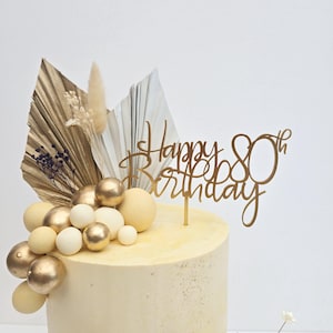 K&ouml;nnte beinhalten: Ein Geburtstagskuchen mit einem goldenen "Happy 80th Birthday" Tortenaufsatz. Der Kuchen ist mit goldenen und wei&szlig;en Kugeln, Trockenblumen und f&auml;cherf&ouml;rmigen Bl&auml;ttern verziert. Der Kuchen hat ein hellgelbes Frosting.