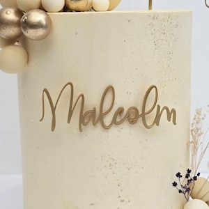 K&ouml;nnte beinhalten: Ein dekorierter Kuchen mit dem Namen "Malcolm" in goldener Schrift. Der Kuchen ist hellbeige mit goldenen Akzenten und mit goldenen und wei&szlig;en dekorativen Kugeln und Trockenblumen verziert. Ein festliches Dessert.