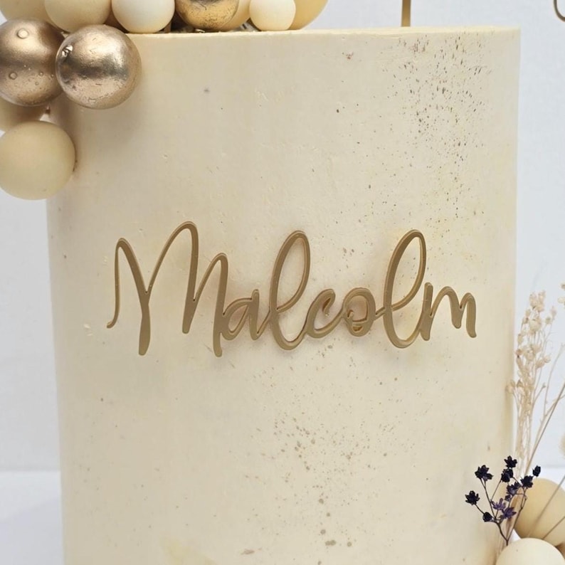 K&ouml;nnte beinhalten: Ein dekorierter Kuchen mit dem Namen "Malcolm" in goldener Schrift. Der Kuchen ist hellbeige mit goldenen Akzenten und mit goldenen und wei&szlig;en dekorativen Kugeln und Trockenblumen verziert. Ein festliches Dessert.