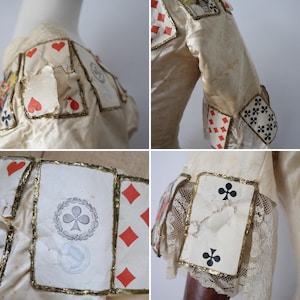 May include: Cream-colored garment adorned with playing card motifs. The cards, featuring hearts, diamonds, clubs, and spades, are outlined with gold trim. The sleeves have lace and card details. The garment appears vintage or antique.