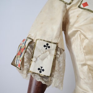 May include: Cream-colored costume with playing card accents. The sleeves feature playing card suits, including clubs, diamonds, and a king. Gold trim and lace details adorn the sleeves. The fabric has a slightly aged appearance.