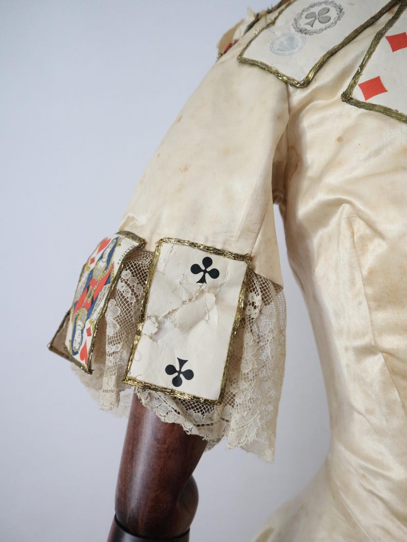 May include: Cream-colored costume with playing card accents. The sleeves feature playing card suits, including clubs, diamonds, and a king. Gold trim and lace details adorn the sleeves. The fabric has a slightly aged appearance.