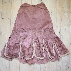 May include: A vintage mauve skirt with a flowing silhouette. The skirt features a textured fabric with a leaf pattern, pleated detailing, and a scalloped hemline trimmed with cream-colored lace. The skirt is displayed on a wooden surface.