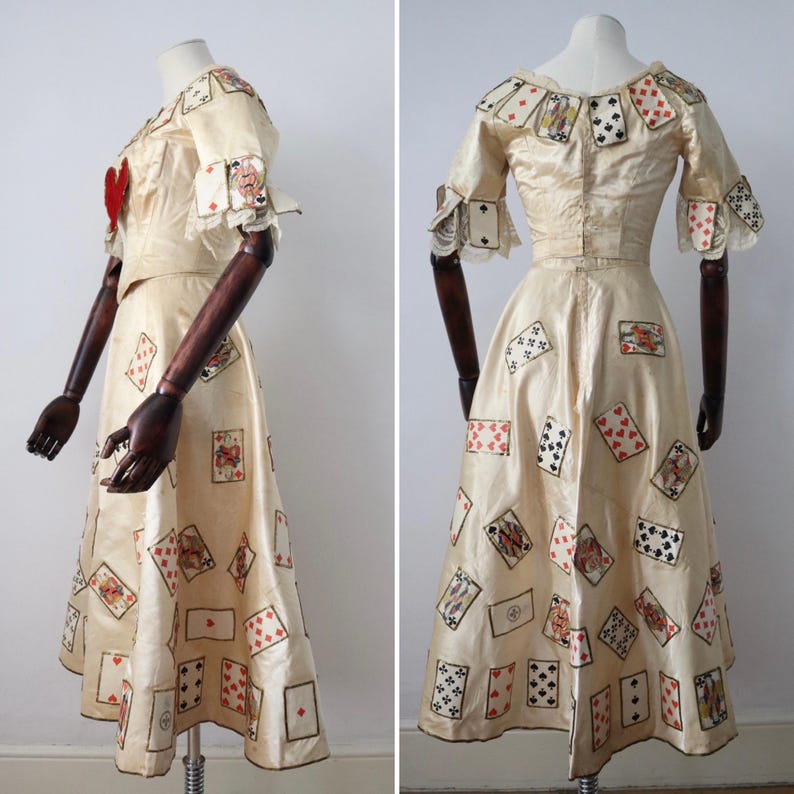 May include: A cream-colored dress adorned with playing card motifs. The dress features short sleeves with ruffled cuffs and a fitted bodice. Playing card designs are scattered across the skirt and neckline, with a red heart accent on the bodice.