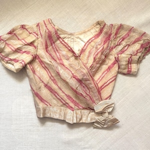 May include: A cream-colored wrap top with pink diagonal stripes. The top features short, puffed sleeves and a bow detail at the waist. The fabric has a slightly sheer, textured appearance, suggesting a vintage or delicate design.