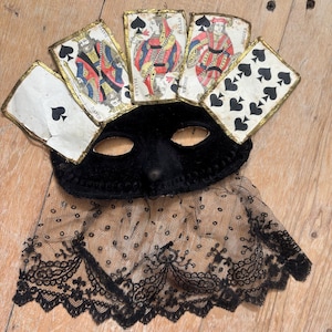 May include: A black velvet masquerade mask with playing cards attached as a crown. The mask has eye holes and a lace trim. The playing cards are antique and feature illustrations of kings and queens.