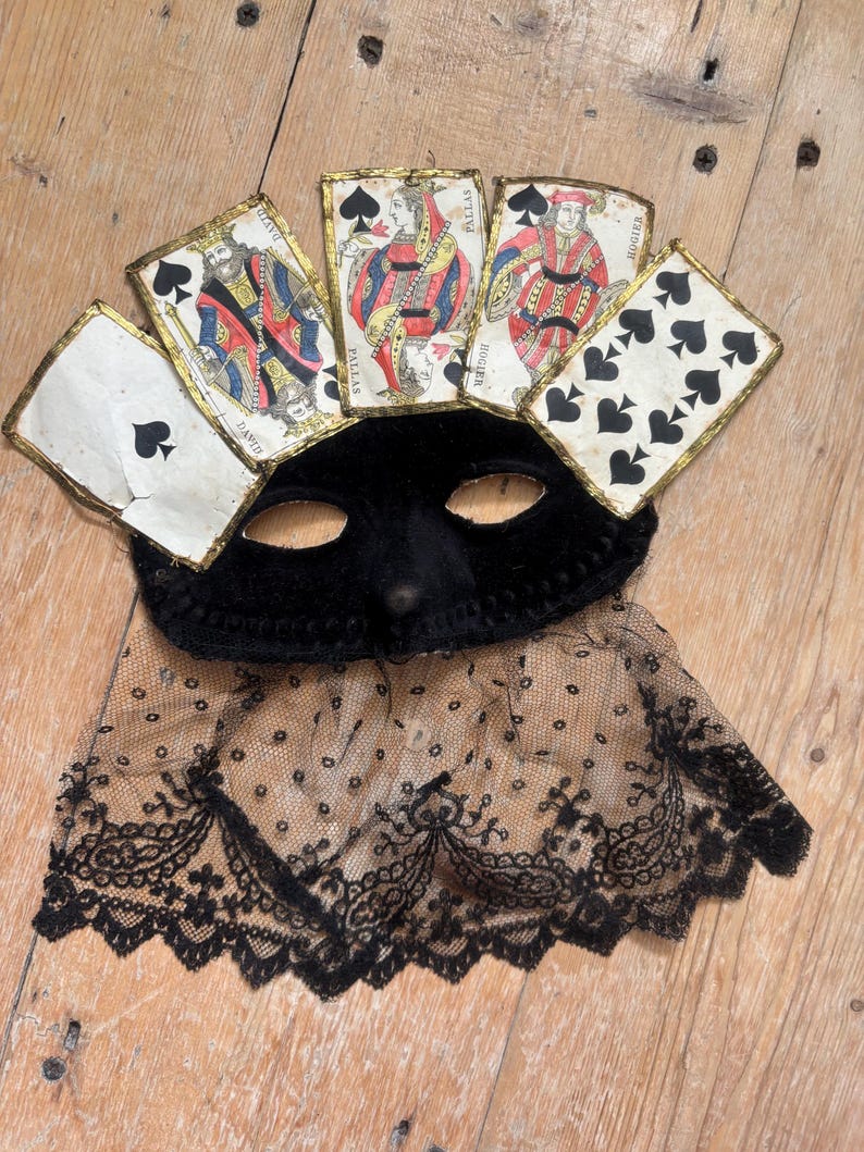May include: A black velvet masquerade mask with playing cards attached as a crown. The mask has eye holes and a lace trim. The playing cards are antique and feature illustrations of kings and queens.