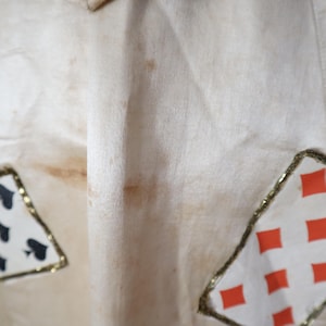 May include: Cream-colored fabric with playing card suit patches. The patches are outlined with gold trim and feature the suits of spades and diamonds. The fabric has some brown staining.