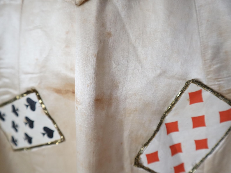 May include: Cream-colored fabric with playing card suit patches. The patches are outlined with gold trim and feature the suits of spades and diamonds. The fabric has some brown staining.