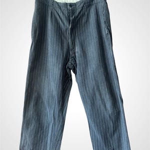 May include: A pair of dark grey trousers featuring a vertical pinstripe pattern. These trousers include a buttoned waistband, a front zip fly, and side pockets. The fabric displays a textured appearance with visible wear near the hemline.